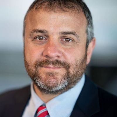 Portrait of a middle-aged man with short brown hair and a salt-and-pepper beard, wearing a suit and red striped tie, looking directly at the camera.